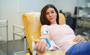 Frau sitzt auf einem gelben Stuhl und spendet Blut, mit einem Schlauch am Arm befestigt.