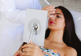 Eine Person liegt auf einem Stuhl und hält einen Spiegel, während eine andere Person in weißer Kleidung ein kosmetisches Gerät auf das Gesicht der liegenden Person richtet.