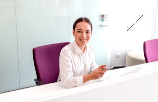 Eine Person sitzt an einem weißen Empfangstresen, hält ein Smartphone und lächelt. Im Hintergrund sind lila Stühle und eine Glaswand zu sehen.