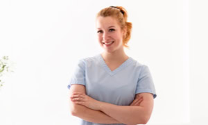 Eine Person in hellblauer medizinischer Kleidung steht mit verschränkten Armen und lächelt.