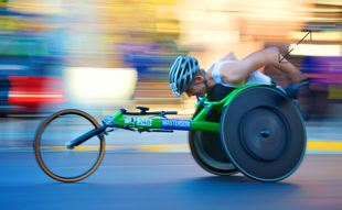 Ein Sportler in einem Rennrollstuhl fährt schnell auf einer Straße, trägt einen Helm und ein ärmelloses Oberteil.
