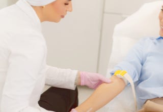 Eine Person in einem weißen Kittel nimmt einer sitzenden Person Blut ab.