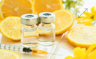 Zwei Glasfl&auml;schchen mit Fl&uuml;ssigkeit, eine Spritze und aufgeschnittene Zitronen auf einem Tisch mit gelben Blumen im Hintergrund.