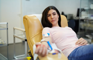 Frau sitzt in einem gelben Stuhl und spendet Blut, mit einer Nadel im Arm und einem Schlauch, der mit einem medizinischen Gerät verbunden ist.