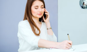 Eine Frau mit langen braunen Haaren telefoniert und schreibt gleichzeitig etwas auf ein Blatt Papier. Sie trägt ein weißes Oberteil und sitzt an einem Tisch.
