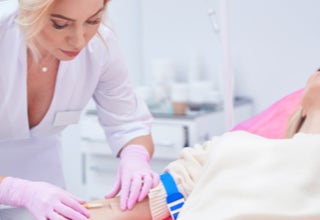 Eine Person in weißer Kleidung und rosa Handschuhen führt eine medizinische Prozedur an einer liegenden Person durch.