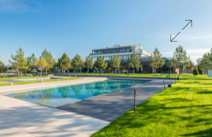 Ein rechteckiger Pool in einem gepflegten Garten mit grünem Rasen, umgeben von Bäumen, im Hintergrund ein großes Gebäude unter blauem Himmel.