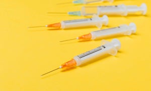 Mehrere Spritzen mit orangefarbenen Kappen liegen auf einem gelben Hintergrund.
