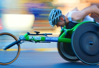 Eine Person in einem Rennrollstuhl mit grünem Rahmen, die einen Helm trägt und sich in Bewegung befindet, mit verschwommenem Hintergrund.