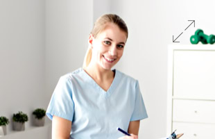 Eine Person in blauer medizinischer Kleidung hält ein Klemmbrett und lächelt in einem hellen Raum mit Pflanzen und einem Regal im Hintergrund.