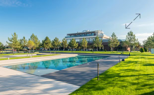 Ein rechteckiger Pool in einem gepflegten Garten mit gr&uuml;nen Rasenfl&auml;chen und B&auml;umen, im Hintergrund ein gro&szlig;es Geb&auml;ude unter blauem Himmel.