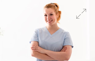 Eine Person in hellblauer medizinischer Kleidung steht mit verschränkten Armen und lächelt vor einem weißen Hintergrund.