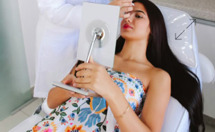 Eine Frau liegt auf einem Stuhl und hält einen Spiegel, während eine Person in weißer Kleidung ihr Gesicht behandelt.