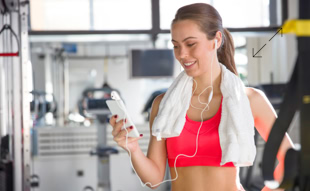 Frau im Fitnessstudio mit Handtuch um den Hals, hält ein Smartphone und trägt Ohrhörer.