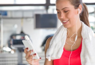 Frau im Fitnessstudio mit Handtuch um den Hals, hält ein Smartphone und trägt Ohrhörer.