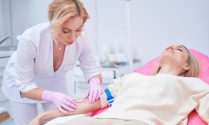 Eine Frau in weißer Kleidung legt einer anderen Frau, die auf einer Liege liegt, eine Infusion an.